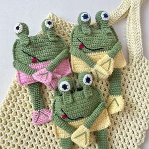 Puede incluir: Tres peluches de rana de crochet en verde, rosa y amarillo. Las ranas tienen ojos grandes y están sentadas en una bolsa de malla de crochet amarilla.