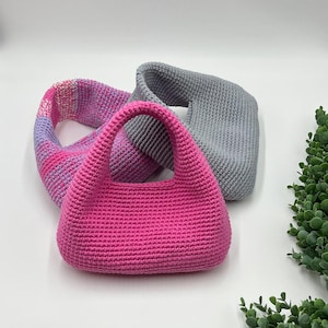 May include: A hot pink crochet handbag with a rounded shape and two handles. A pink, purple, and white striped crochet cowl and a gray crochet cowl are behind the handbag. The items are on a white surface with green foliage in the corner.