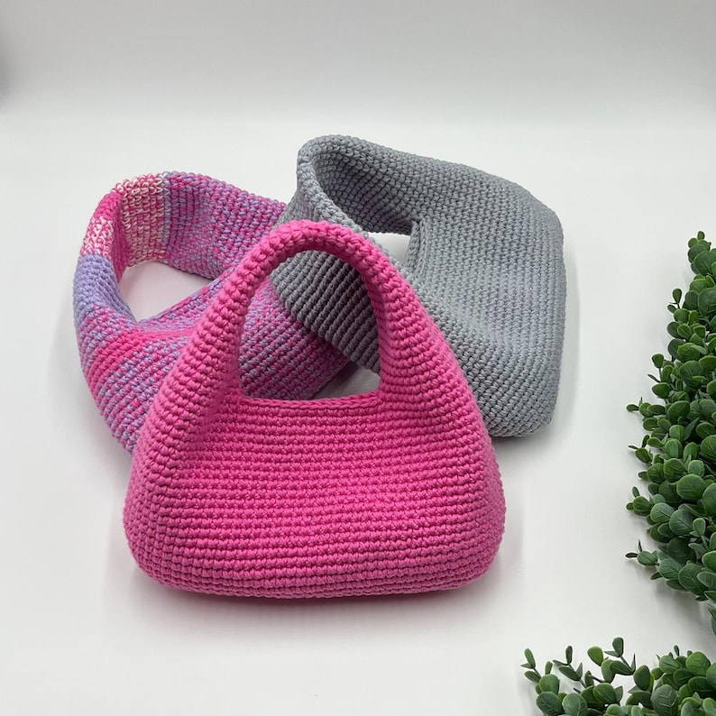 May include: A hot pink crochet handbag with a rounded shape and two handles. A pink, purple, and white striped crochet cowl and a gray crochet cowl are behind the handbag. The items are on a white surface with green foliage in the corner.