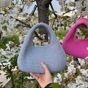 May include: A light blue crochet handbag with a rounded shape and a sturdy handle. The bag is held in front of a background of white flowers and tree branches. A pink crochet bag is also visible.