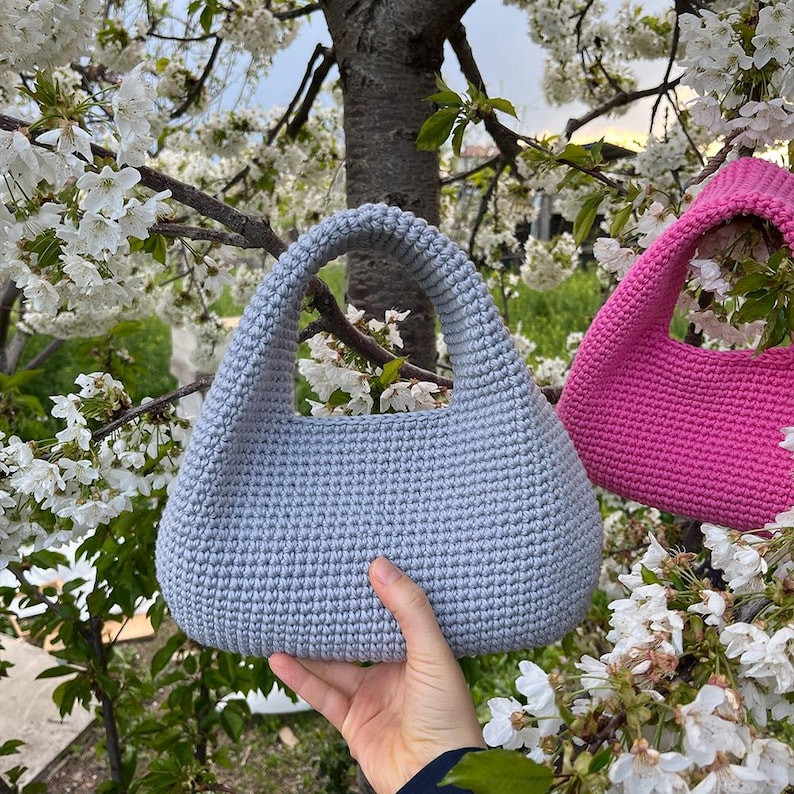 May include: A light blue crochet handbag with a rounded shape and a sturdy handle. The bag is held in front of a background of white flowers and tree branches. A pink crochet bag is also visible.