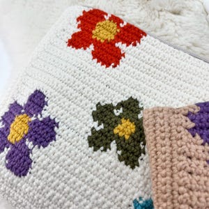 May include: A white crochet cushion featuring vibrant floral patterns. The flowers are red, purple, green, and tan with yellow centres. The cushion has a zip closure and is made from soft, textured yarn.