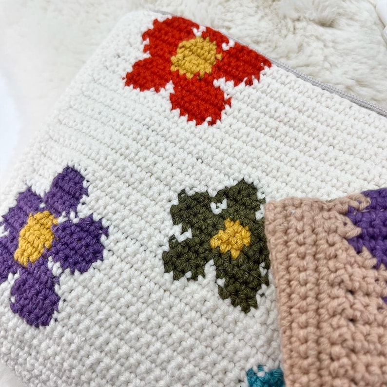 May include: A white crochet cushion featuring vibrant floral patterns. The flowers are red, purple, green, and tan with yellow centres. The cushion has a zip closure and is made from soft, textured yarn.