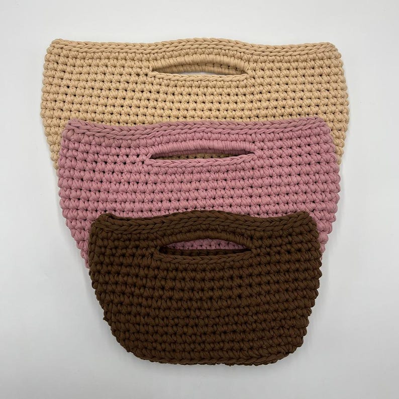 May include: Three stacked crochet handbags with integrated handles. The top bag is beige, the middle bag is dusty rose, and the bottom bag is dark brown. Each bag features a consistent, dense, textured stitch pattern throughout the exterior.