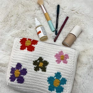 May include: A white crocheted cosmetic bag with colourful flower designs. The bag is surrounded by makeup items, including a brush, eyeliner, lip liner, foundation, and a skincare bottle.