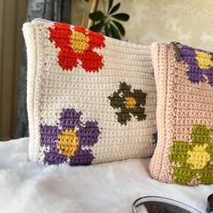 May include: Two crochet pouches with floral designs. One is off-white with red, purple, and olive green flowers. The other is light pink with purple and green flowers. Both have zippers.