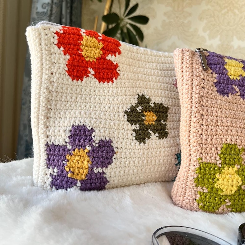 May include: Two crochet pouches with floral designs. One is off-white with red, purple, and olive green flowers. The other is light pink with purple and green flowers. Both have zippers.