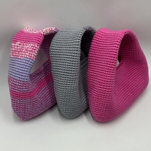 May include: Three handmade crochet bags in varying shades of pink and gray. The bag on the left has a gradient of pink and purple, the middle bag is gray, and the bag on the right is a solid bright pink. The bags are all the same shape.