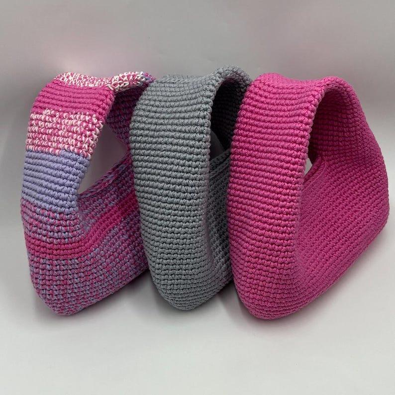 May include: Three handmade crochet bags in varying shades of pink and gray. The bag on the left has a gradient of pink and purple, the middle bag is gray, and the bag on the right is a solid bright pink. The bags are all the same shape.