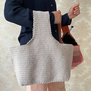 May include: A large, light beige crochet tote bag with a wide handle. The bag is held by a person wearing a navy blue shirt and pink pants. A smaller, ombre-colored crochet bag is also visible.