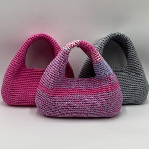 May include: Three crochet handbags in shades of pink and gray. The bags have a rounded shape with a single handle. The central bag features a gradient of pink and purple hues, while the others are solid pink and gray.