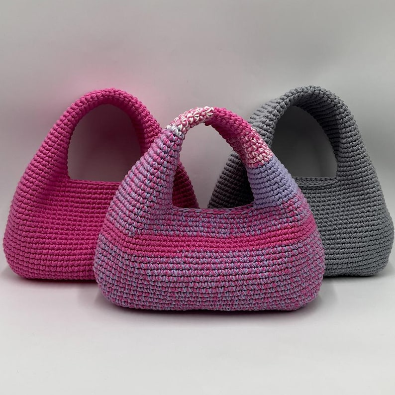 May include: Three crochet handbags in shades of pink and gray. The bags have a rounded shape with a single handle. The central bag features a gradient of pink and purple hues, while the others are solid pink and gray.