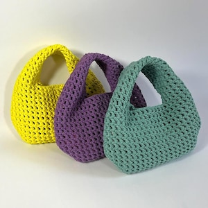 Puede incluir: Tres bolsos pequeños hechos de cordón grueso anudado en amarillo brillante, morado vibrante y verde salvia suave. Cada bolso cuenta con un asa integrada única y una textura de tejido abierto consistente, dispuestos en fila sobre un fondo liso.
