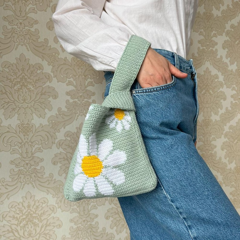 May include: A light green crocheted tote bag with white and yellow daisy flowers. The bag has a knotted handle.