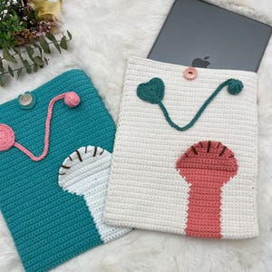 Puede incluir: Dos fundas para tablet de crochet, una turquesa con un diseño rosa y verde y la otra blanca con un diseño rosa y verde.