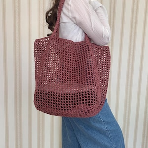 May include: A dusky pink open-weave tote bag with a rectangular shape and strong handles. The bag is made of a crocheted material, creating a net-like appearance. The bag is being carried by a person wearing a white shirt and blue jeans.