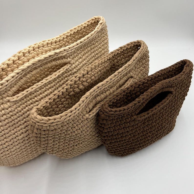 May include: Three handmade crochet tote bags in varying sizes and shades of brown. The largest bag is a light beige, the middle bag is a medium tan, and the smallest is a dark brown. Each bag has a sturdy, braided handle.