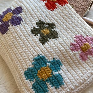 May include: Cream-coloured crocheted blanket with a repeating floral pattern. The flowers are in various colours, including purple, teal, pink, green, and orange, each with a yellow centre. The blanket has a ribbed texture.