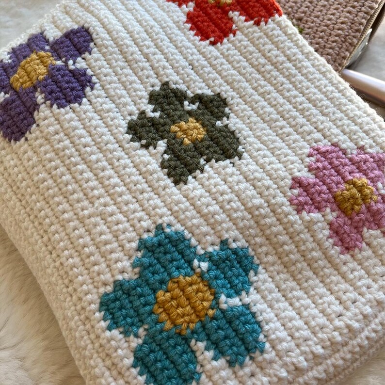 May include: Cream-coloured crocheted blanket with a repeating floral pattern. The flowers are in various colours, including purple, teal, pink, green, and orange, each with a yellow centre. The blanket has a ribbed texture.
