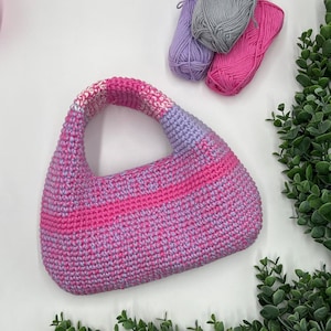 Puede incluir: Bolso de mano de ganchillo hecho a mano en tonos rosa y lavanda. El bolso tiene una forma redondeada con un asa. Tres madejas de hilo en colores coordinados son visibles en el fondo, junto con vegetación.