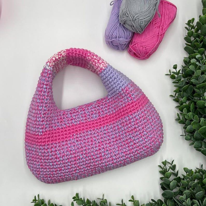 May include: A handmade crochet handbag in shades of pink and lavender. The bag features a rounded shape with a single handle. Three skeins of yarn in coordinating colors are visible in the background, along with greenery.