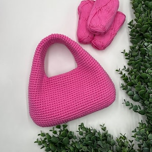 May include: A bright pink, crocheted handbag with a curved handle. Three balls of matching pink yarn are arranged above the bag. Green leafy sprigs are in the background, creating a contrast with the pink items.