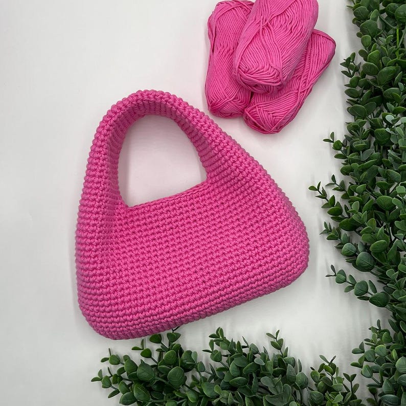 May include: A bright pink, crocheted handbag with a curved handle. Three balls of matching pink yarn are arranged above the bag. Green leafy sprigs are in the background, creating a contrast with the pink items.