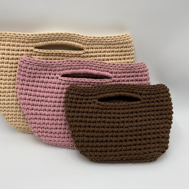 May include: Three handmade crochet bags in varying sizes and colors. The largest bag is a light beige, the middle bag is pink, and the smallest is brown. Each bag has a handle opening at the top, and the bags are made of a thick, textured yarn.