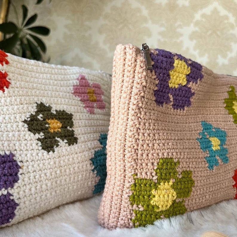 May include: Two crochet pouches with floral patterns. One is off-white with red, pink, green, and purple flowers. The other is light pink with purple, green, blue, and yellow flowers. Both have a zip closure.