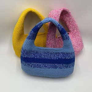 Op de afbeelding: Drie handgemaakte gehaakte handtassen in verschillende kleuren. Eén tas is effen geel, één is gemêleerd roze en de voorste tas heeft horizontale strepen in tinten lichtblauw, middenblauw en donkerblauw met een geïntegreerd handvat.