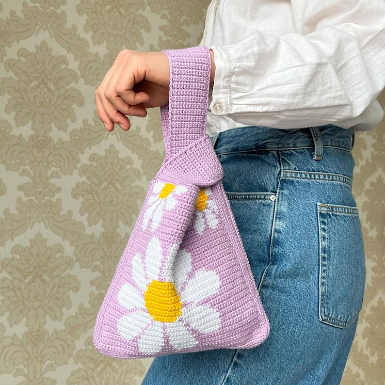 May include: A lavender crocheted tote bag with a knotted handle and white daisy flower designs. The bag has yellow centers on the daisies.