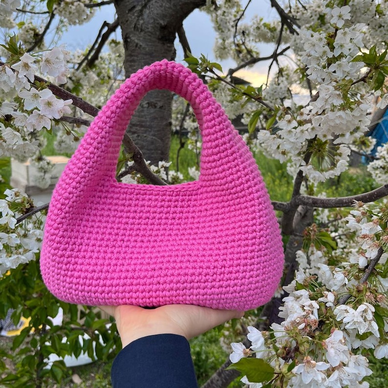 May include: A bright pink, handmade crochet handbag with a rounded shape and a single handle. The bag is made of thick yarn, and the texture is visible. The background features white flowers and tree branches.