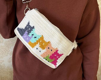Op de afbeelding: Een witte gehaakte crossbodytas met een kleurrijk kattenontwerp. De tas heeft een ritssluiting en een verstelbare riem. Het kattenontwerp bevat tinten paars, blauwgroen, geel, perzik en roze.