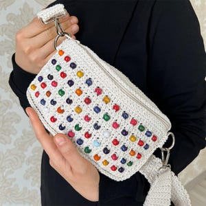 Könnte beinhalten: Eine weiße gehäkelte Tasche mit Reißverschluss und einem langen Riemen. Die Tasche ist mit bunten Holzperlen verziert.