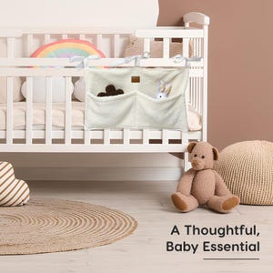 May include: A cream-colored baby crib organizer with two pockets, hanging from the crib. The pockets contain a brown bear and a white bunny. The text "A Thoughtful, Baby Essential" is at the bottom of the image.