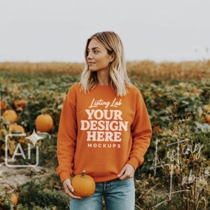 Könnte beinhalten: Ein orangefarbenes Langarm-Sweatshirt mit dem weißen Schriftzug Listing Lab YOUR DESIGN HERE MOCKUPS auf der Vorderseite. Das Kleidungsstück hat einen Rundhalsausschnitt sowie gerippte Bündchen und wird vor einem Kürbisfeld gehalten.