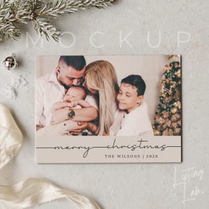 May include: A rectangular holiday greeting card featuring a photograph of a family of four, including a baby, with the text "merry christmas" in a cursive script and "THE WILSONS | 2026" printed in a clean, sans-serif font on a light beige base.