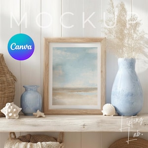 May include: A framed landscape art print with a light wood frame sits on a rustic shelf, alongside a blue vase with dried flowers and decorative seashells. The art print features a beach scene with a blue sky and sandy shore. The Canva logo is visible.