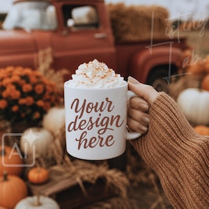 May include: A white ceramic mug filled with whipped cream topped with cinnamon, featuring the brown text Your design here printed on the front. The mug is held by a hand wearing a brown knitted sweater sleeve against a rustic autumn background.