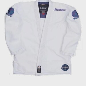 May include: A white martial arts gi jacket featuring purple and blue circular patches on both shoulders. The chest displays the text SHOYOROLL in purple, while the inner collar label reads CLASSIC. A small circular logo is on the lower left.