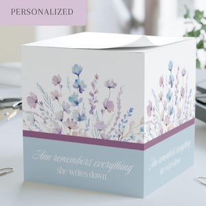 May include: A square paper memo cube featuring a watercolor floral design in shades of blue and purple on a white background. The base is a solid light blue with a dark purple stripe and the white text: Ann remembers everything she writes down.