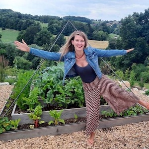 Op de afbeelding: Een vrouw in een blauw denim jack en een gepatroneerde broek staat in een verhoogd tuinbed. Het tuinbed is gemaakt van hout en heeft meerdere niveaus. De vrouw glimlacht en heeft haar armen uitgestrekt.
