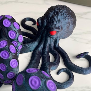 May include: A 3D printed octopus figurine with a dark blue body and purple suction cups. The octopus has red eyes and is posed with its tentacles curled.
