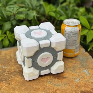 May include: A white, grey, and pink cube-shaped object with a heart design, similar to a video game prop. The cube is on a brown surface, with a yellow can in the background.