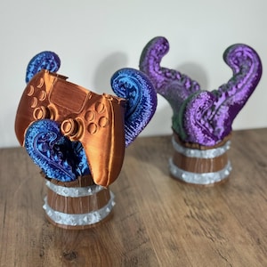 May include: Two decorative gaming controller holders. One holder is holding a copper-colored controller with blue and purple tentacles. The other holder has purple tentacles. Both holders are in a wooden barrel design with silver accents.
