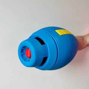 May include: A blue and yellow plastic toy gun with a red button on the front. The gun is designed to look like a futuristic weapon.