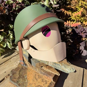 Peut inclure: Un casque vert avec une sangle marron et un masque facial beige. Le masque a un œil en maille rose et est monté sur une base en pierre. Le casque est de style militaire.
