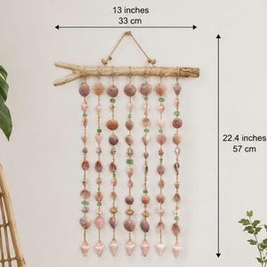 Può includere: Una decorazione da parete composta da un pezzo orizzontale di legno galleggiante con otto fili verticali di conchiglie assortite e piccole perline verdi sospese. L'oggetto misura 33 cm di larghezza e 57 cm di altezza totale dall'alto al basso.