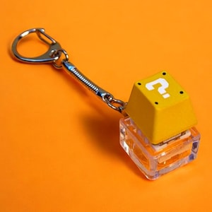 Puede incluir: Un llavero de bloque de interrogación amarillo con una caja de plástico transparente. El llavero está unido a una cadena de metal plateado.