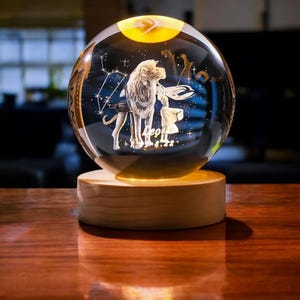 Può includere: Una sfera di vetro trasparente con una base in legno, illuminata dall'interno. La sfera presenta un design inciso in 3D del segno zodiacale Leone, che include un leone e stelle. Il testo "Leo 8.22" è anche inciso sulla sfera.
